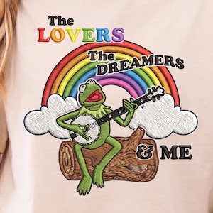 May include: Embroidered design featuring Kermit the Frog playing a banjo, sitting on a log under a rainbow. Text reads "The Lovers, The Dreamers & Me" in colorful letters. The design is on a light beige fabric.