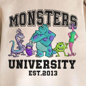 May include: A beige sweatshirt featuring the text "MONSTERS UNIVERSITY" in black, with the year "EST.2013" below. The design includes embroidered cartoon characters in various colors, including blue, green, purple, and pink.