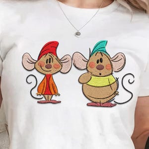 Könnte beinhalten: Weißes T-Shirt mit einem gestickten Design von zwei Zeichentrickmäusen. Eine Maus trägt einen roten Hut und einen orangefarbenen Mantel, die andere einen grünen Hut und ein gelbes Hemd. Die Mäuse haben große Ohren und lächeln. Das Design ist auf dem T-Shirt zentriert.