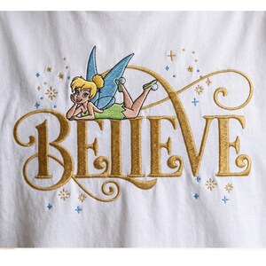 May include: White t-shirt featuring gold embroidered text spelling "BELIEVE". A cartoon fairy with blonde hair, blue wings, and a green dress is embroidered above the word. Blue and gold accents surround the text.