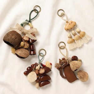 May include: Four handmade keychains with natural elements. Each keychain features a metal ring, a loop of cord, and various organic materials like nuts, seeds, and wood. One has a wooden cloud charm and tassel, another has a bow.