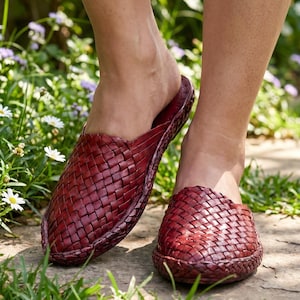 Könnte beinhalten: Ein Paar dunkelrote Slipper mit einem gewebten Lederobermaterial. Die Schuhe haben eine geschlossene Zehenpartie mit abgerundeter Vorderseite und ein geflochtenes Zierdetail entlang der Sohlenkante, was für eine strukturierte Optik sorgt.
