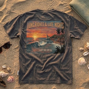 Puede incluir: Camiseta gris oscuro con un diseño de puesta de sol en la playa. El diseño incluye el texto "LONG RIGHTS & LATE NIGHTS" y "GLORY DAZE SURF CO". Gafas de sol, conchas marinas y una toalla sobre la arena.