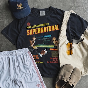 May include: A navy blue baseball cap with a Cadillac logo, a black t-shirt featuring Supernatural graphics and text, white athletic shorts with red side stripes, a beige canvas tote bag, yellow-tinted sunglasses, and tan suede slip-on shoes.