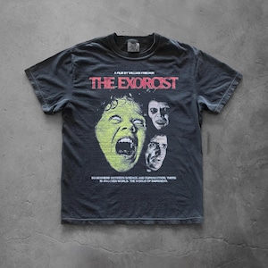 Pepper Comfort Colors t-shirt featuring The Exorcist graphic print, displayed flat on a concrete background. The design shows a vintage horror movie collage with bold red title text, eerie character portraits, and distressed retro-style artwork.