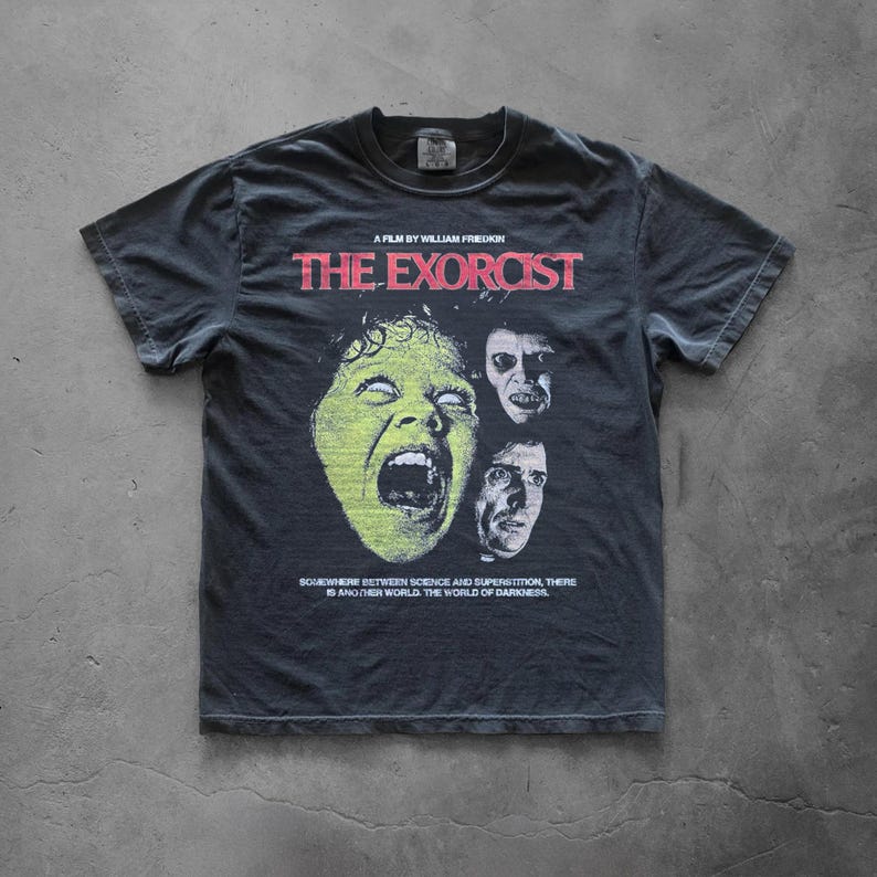 Pepper Comfort Colors t-shirt featuring The Exorcist graphic print, displayed flat on a concrete background. The design shows a vintage horror movie collage with bold red title text, eerie character portraits, and distressed retro-style artwork.