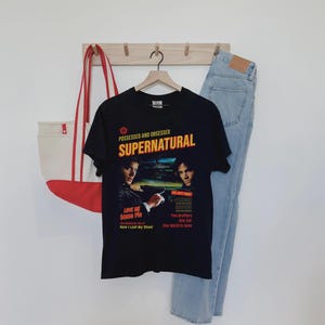 May include: A black short-sleeve t-shirt featuring a graphic print with the text "POSSESSED AND OBSESSED SUPERNATURAL" and images of two men. The print includes additional text: "LOVE ME Some Pie", "How I Lost My Shoe!", "Two Brothers One Car One World to Save".