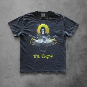 Puede incluir: Camiseta gris oscuro con un gráfico de una figura con los brazos extendidos, un halo amarillo y un diseño de pájaro. El texto "The Crow" está impreso en amarillo debajo de la imagen. La camiseta es de material suave.