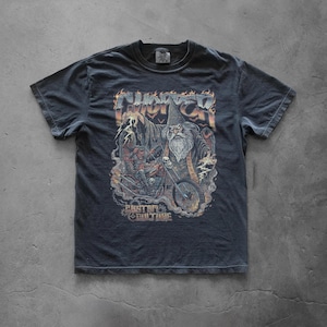 Pepper vintage-style graphic T-shirt laid flat on a concrete surface featuring a bold fantasy biker illustration with a wizard casting lightning, skull details, and a chopper motorcycle in a distressed heavy metal art style.