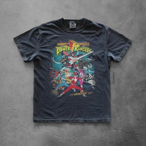 Flat lay of a charcoal gray Comfort Colors t-shirt on a concrete floor. The front features a large, colorful Power Rangers "It's Morphin' Time" graphic. Shows the boxy, vintage-wash fit and durable ribbed collar of the heavyweight tee.
