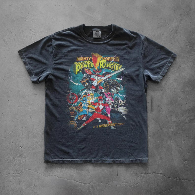 Flat lay of a charcoal gray Comfort Colors t-shirt on a concrete floor. The front features a large, colorful Power Rangers "It's Morphin' Time" graphic. Shows the boxy, vintage-wash fit and durable ribbed collar of the heavyweight tee.