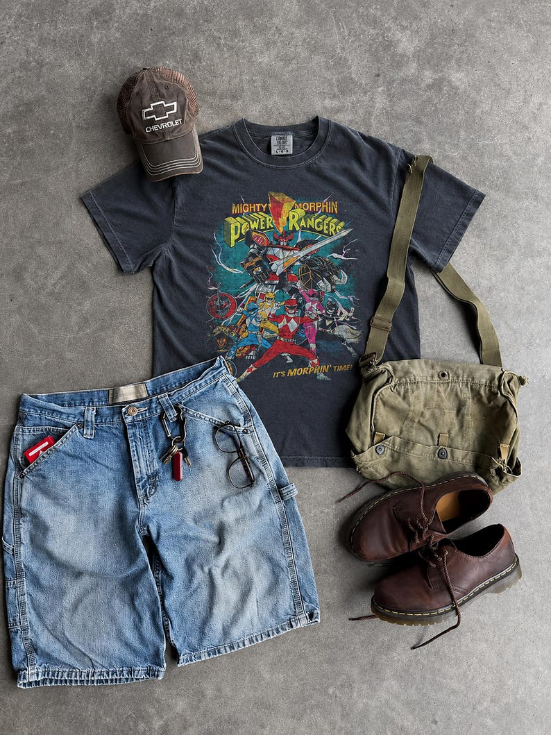 K&ouml;nnte beinhalten: Eine Zusammenstellung l&auml;ssiger Kleidung, darunter ein graues T-Shirt mit einem "Mighty Morphin Power Rangers" Aufdruck, blaue Jeansshorts, eine braune Baseballkappe, eine gr&uuml;ne Canvas-Tasche und braune Lederschuhe.
