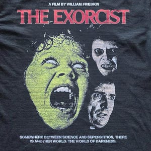 Black Comfort Colors t-shirt with The Exorcist graphic print, shown flat on a concrete background. The design features a retro horror movie collage with bold red lettering, unsettling character artwork, and classic poster-inspired details.