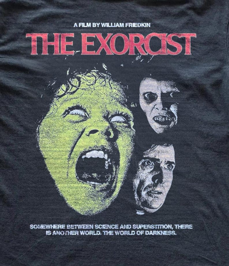 Black Comfort Colors t-shirt with The Exorcist graphic print, shown flat on a concrete background. The design features a retro horror movie collage with bold red lettering, unsettling character artwork, and classic poster-inspired details.
