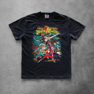 A black graphic t-shirt laid flat on a gray concrete background. The center showcases a distressed retro Power Rangers design with bold yellow typography. The high-quality cotton fabric and classic streetwear silhouette are clearly visible.