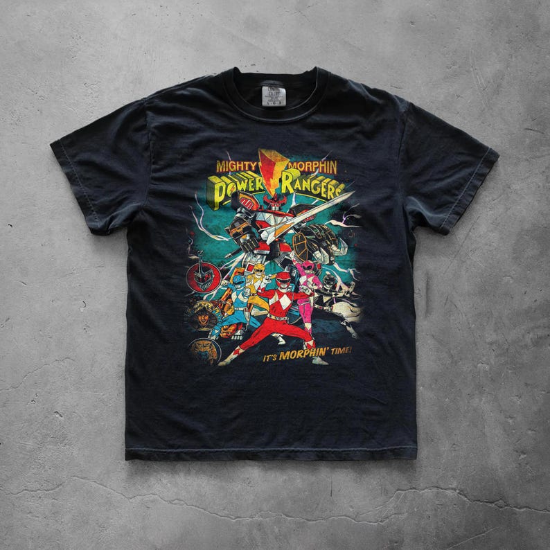 A black graphic t-shirt laid flat on a gray concrete background. The center showcases a distressed retro Power Rangers design with bold yellow typography. The high-quality cotton fabric and classic streetwear silhouette are clearly visible.