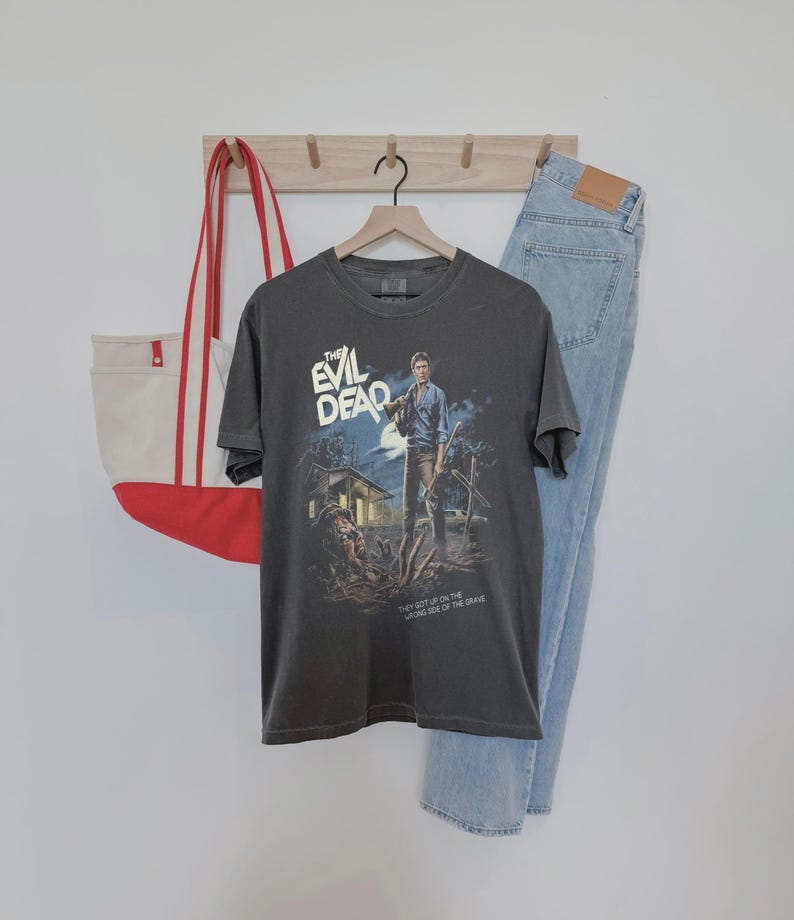 Puede incluir: Una camiseta gris oscuro con un dise&ntilde;o de cartel de pel&iacute;cula de "The Evil Dead". El gr&aacute;fico muestra a un hombre con un arma frente a una casa, con el texto "THE EVIL DEAD" y "THEY GOT UP ON THE WRONG SIDE OF THE GRAVE."