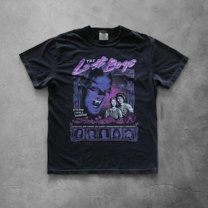 Black graphic T-shirt displayed flat on a concrete surface showcasing a large retro The Lost Boys movie-inspired print with neon purple and blue horror-themed artwork.