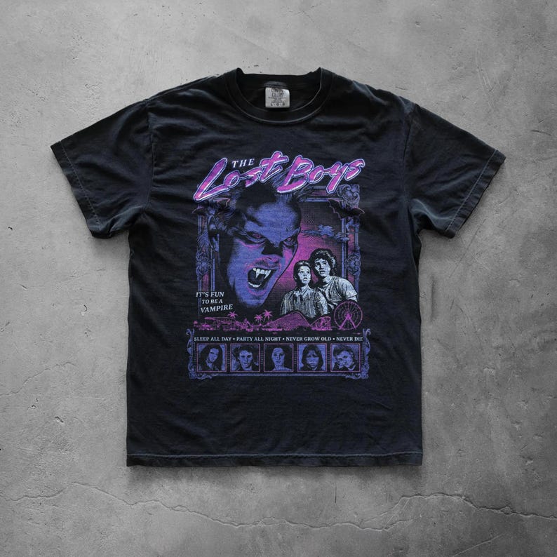 Black graphic T-shirt displayed flat on a concrete surface showcasing a large retro The Lost Boys movie-inspired print with neon purple and blue horror-themed artwork.