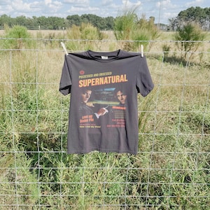 May include: A charcoal gray short-sleeve t-shirt featuring a graphic print with the text "POSSESSED AND OBSESSED SUPERNATURAL" and images of two individuals. The print includes additional text such as "LOVE ME SOME PIE" and "Two Brothers".