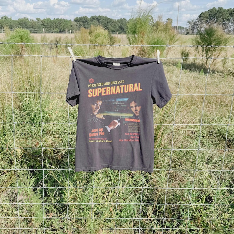 May include: A charcoal gray short-sleeve t-shirt featuring a graphic print with the text "POSSESSED AND OBSESSED SUPERNATURAL" and images of two individuals. The print includes additional text such as "LOVE ME SOME PIE" and "Two Brothers".