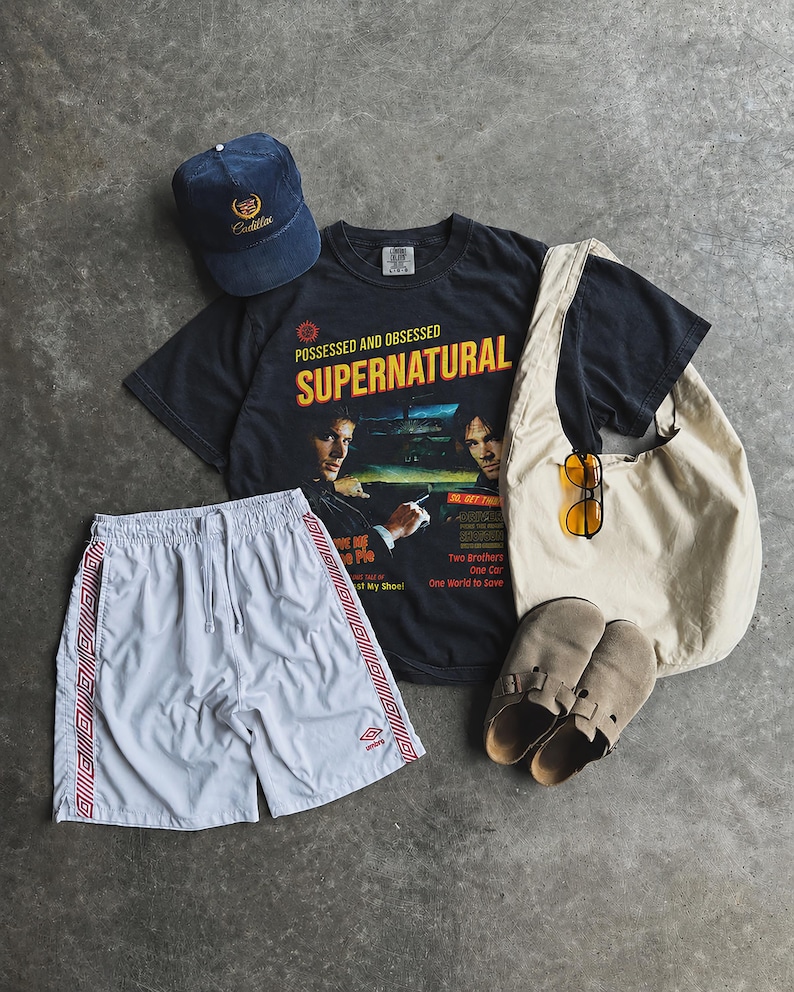 May include: A navy blue baseball cap with a Cadillac logo, a black t-shirt featuring Supernatural graphics and text, white athletic shorts with red side stripes, a beige canvas tote bag, yellow-tinted sunglasses, and tan suede slip-on shoes.