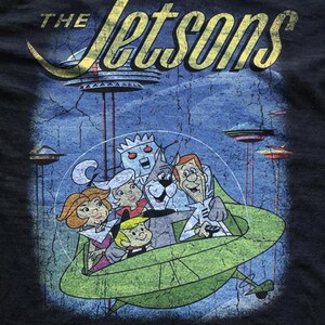 May include: Black t-shirt featuring a distressed graphic of the Jetsons family in a green spaceship. The text "The Jetsons" is in yellow, with a retro space-age design. The background shows flying saucers and a blue sky.