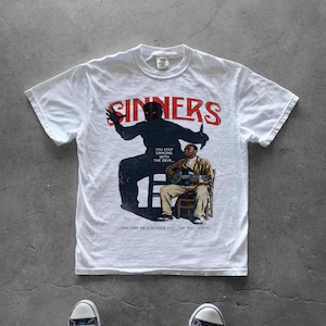 Może przedstawiać: Biały t-shirt z krótkim rękawem z nadrukiem graficznym i czerwonym napisem SINNERS u góry. Wzór przedstawia ciemną sylwetkę postaci górującej nad osobą grającą na gitarze na krześle, z tekstem YOU KEEP DANCING WITH THE DEVIL... ONE DAY HE'S GONNA FOLLOW YOU HOME!