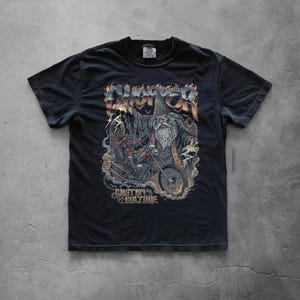 Black graphic T-shirt displayed flat on a concrete floor showcasing a large distressed wizard biker illustration with skulls, flames, and custom culture typography in a retro heavy metal style.