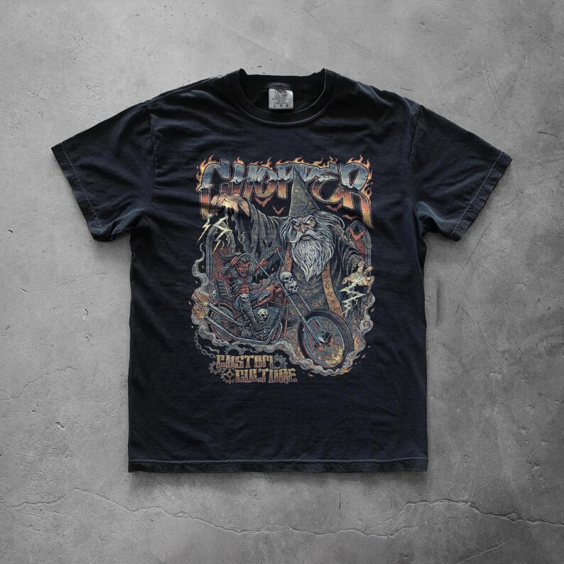 Black graphic T-shirt displayed flat on a concrete floor showcasing a large distressed wizard biker illustration with skulls, flames, and custom culture typography in a retro heavy metal style.