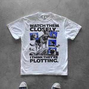 Puede incluir: Camiseta blanca con un diseño gráfico de perros y el texto "WATCH THEM CLOSELY... I THINK THEY'RE PLOTTING." El diseño incluye un pastor alemán, un beagle, un pug y otros perros.