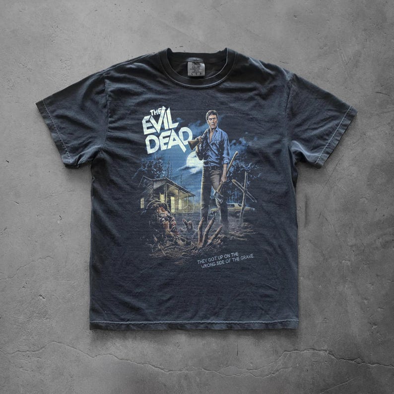 A faded dark grey vintage-style t-shirt laid flat on a concrete surface. The shirt features a large graphic print for the classic horror movie "The Evil Dead". The design illustrates the character Ash Williams holding a shotgun and an axe, a creepy cabin in the woods, a classic car, and a screaming deadite emerging from a grave. The movie title is at the top left, and the tagline "THEY GOT UP ON THE WRONG SIDE OF THE GRAVE." is printed at the bottom right.