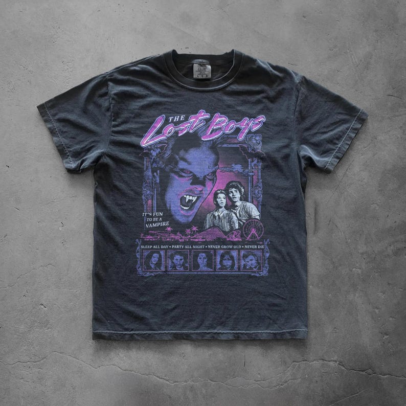Ivoly vintage-style graphic T-shirt laid flat on a concrete floor featuring a bold retro The Lost Boys horror design with neon purple and blue tones and distressed 80s movie-style artwork.