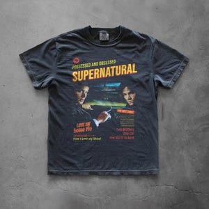 May include: A charcoal gray short-sleeve t-shirt featuring a graphic print with the text SUPERNATURAL, POSSESSED AND OBSESSED, LOVE ME SOME PIE, HOW I LOST MY SHOE, SO GET THIS, DRIVER PICKS THE MUSIC, SHOTGUN SHUTS HIS CAKEHOLE, TWO BROTHERS, ONE CAR, ONE WORLD TO SAVE.