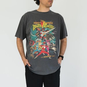 K&ouml;nnte beinhalten: Ein anthrazitfarbenes T-Shirt mit einer farbenfrohen Grafik der Mighty Morphin Power Rangers. Das Design enth&auml;lt den Text "Mighty Morphin Power Rangers" und "It's Morphin' Time!". Das Shirt hat einen Vintage-Stil und ein abgenutztes Aussehen.