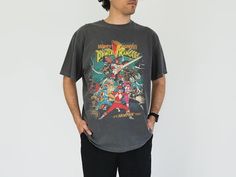 K&ouml;nnte beinhalten: Ein anthrazitfarbenes T-Shirt mit einer farbenfrohen Grafik der Mighty Morphin Power Rangers. Das Design enth&auml;lt den Text "Mighty Morphin Power Rangers" und "It's Morphin' Time!". Das Shirt hat einen Vintage-Stil und ein abgenutztes Aussehen.
