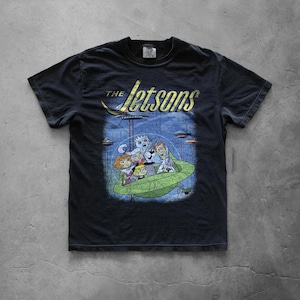 May include: Black t-shirt featuring a graphic of The Jetsons cartoon family in a green spaceship. The shirt has the show's logo in gold lettering above the image. The design is distressed for a vintage look.