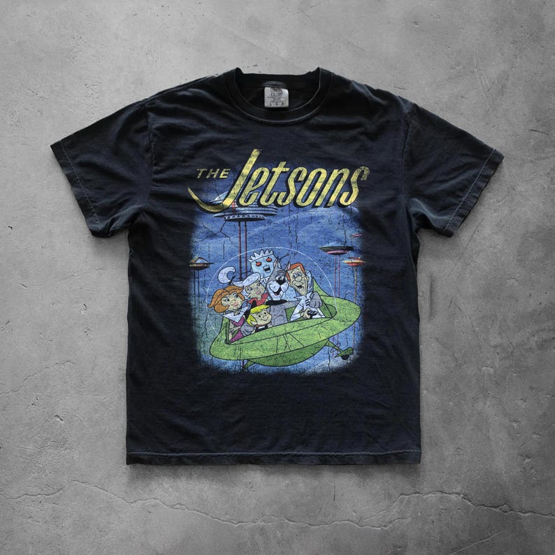 May include: Black t-shirt featuring a graphic of The Jetsons cartoon family in a green spaceship. The shirt has the show's logo in gold lettering above the image. The design is distressed for a vintage look.