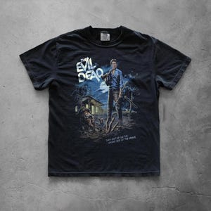 A black vintage-style short-sleeve t-shirt laid flat on a grey concrete floor. It features the "The Evil Dead" movie poster graphic, showing Ash Williams armed with a shotgun and an axe standing in a spooky graveyard setting with a cabin, an old car, and a zombie rising from the ground under a full moon.