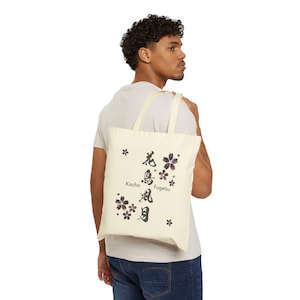 May include: Cream-colored tote bag with black and purple floral design and Japanese text. The bag is carried over the shoulder and features the words "Kacho Fugetsu". The tote bag is made of a sturdy canvas material.