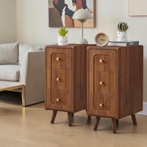 May include: Two matching wooden bedside tables, each featuring three drawers with small gold-toned circular knobs. The cabinets have a warm brown wood grain finish, rounded vertical edges, and are supported by four tapered wooden legs.