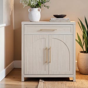 May include: A light-colored wooden cabinet with a drawer and two doors. The cabinet has gold-colored handles and a curved design on the doors. A white vase with flowers, a black bowl, and books are on top. A potted plant is to the right.