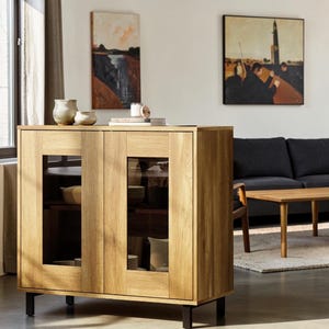 May include: A light oak wood sideboard cabinet featuring two doors with rectangular glass panels, black metal legs, and internal shelving. The cabinet is rectangular in shape with a visible wood grain finish and a clean, structured design.