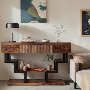 May include: A console table featuring a dark wood grain finish with a central drawer that has a vertical slatted front. The table is supported by a black metal frame with a geometric, stepped design that creates two lower shelves for storage.