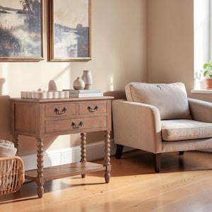 May include: A light brown wooden console table with three drawers, featuring dark metal pulls and turned spindle legs. The table includes a lower shelf and is topped with two ceramic vases and a stack of books, positioned next to an armchair.