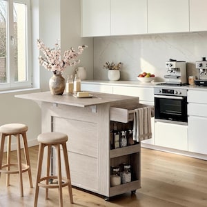 May include: A light wood kitchen island on wheels with a drop-leaf top, two open storage shelves, and a side towel rack. Two matching light wood bar stools with beige upholstered seats are positioned in front of the island.