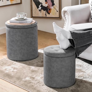 May include: Two gray storage ottomans with a tufted top and a wooden lid. One ottoman has a cup and books on top. The ottomans are cylindrical and made of a textured material. They are placed on a rug.
