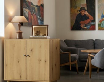 May include: A light oak wood cabinet with two doors and small silver knobs, featuring a visible wood grain pattern. The cabinet stands on a sturdy base and is topped with a wooden table lamp and a framed picture.