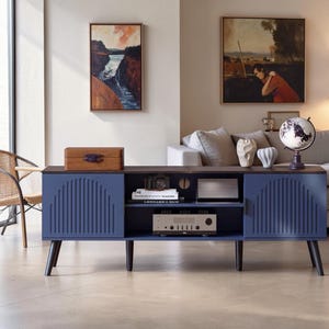 May include: A long, blue media console with a dark wood top and four tapered legs. The unit features two side cabinets with vertical ribbed door panels, an open center shelf containing a camera, books, and a silver audio receiver.