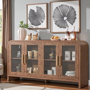 May include: A wooden sideboard with glass doors and gold handles. The cabinet is filled with various items, including coffee containers, bottles, and decorative objects. Two framed black and white art pieces hang above.